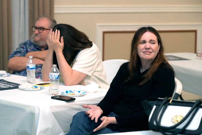 Three people sitting at a table; one man wears glasses, a woman covers her face, and another woman looks ahead with a puzzled expression. Names unknown