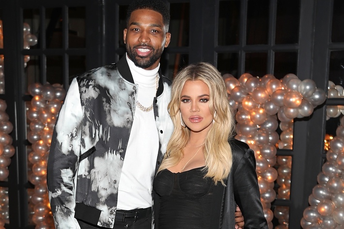 Khloé Kardashian in a fitted dress and Tristan Thompson in a patterned jacket pose together at an event with a balloon backdrop