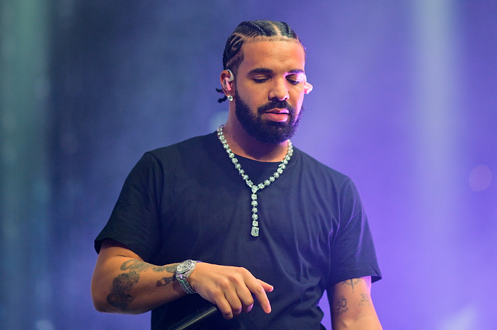 Drake on stage performing, wearing a black T-shirt, diamond necklace, and a watch, with braided hair styled back