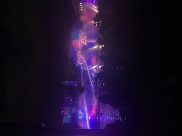 Water show in progress at night, with colorful lights and projections on water sprays. A crowd of people watch and capture the event with their cameras