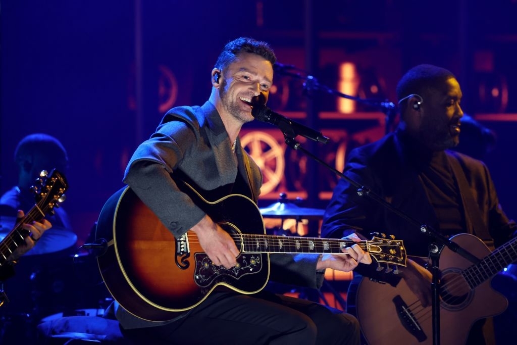 Justin Timberlake plays an acoustic guitar and sings into a microphone on stage, accompanied by another musician on a guitar