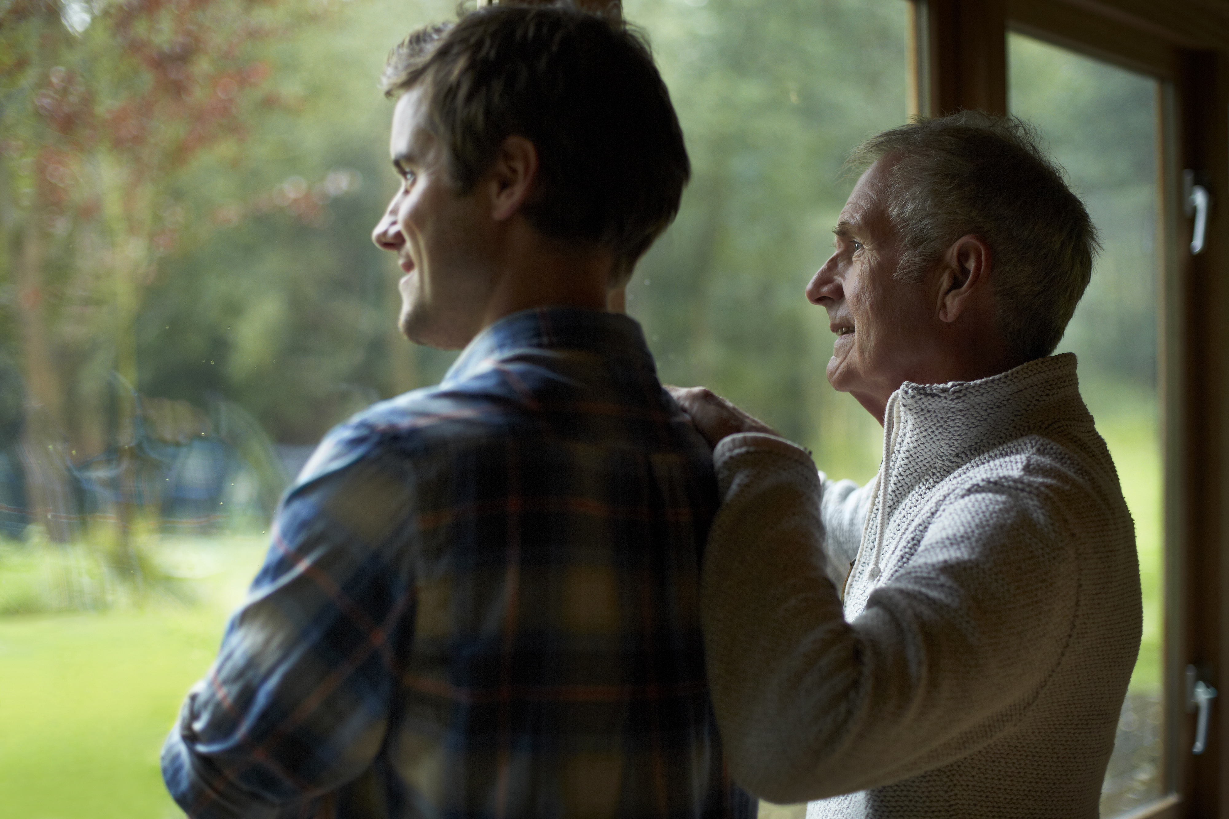 Two people stand together, one younger and one older, looking out a window with the older person resting their hand on the younger person's shoulder. Names unknown
