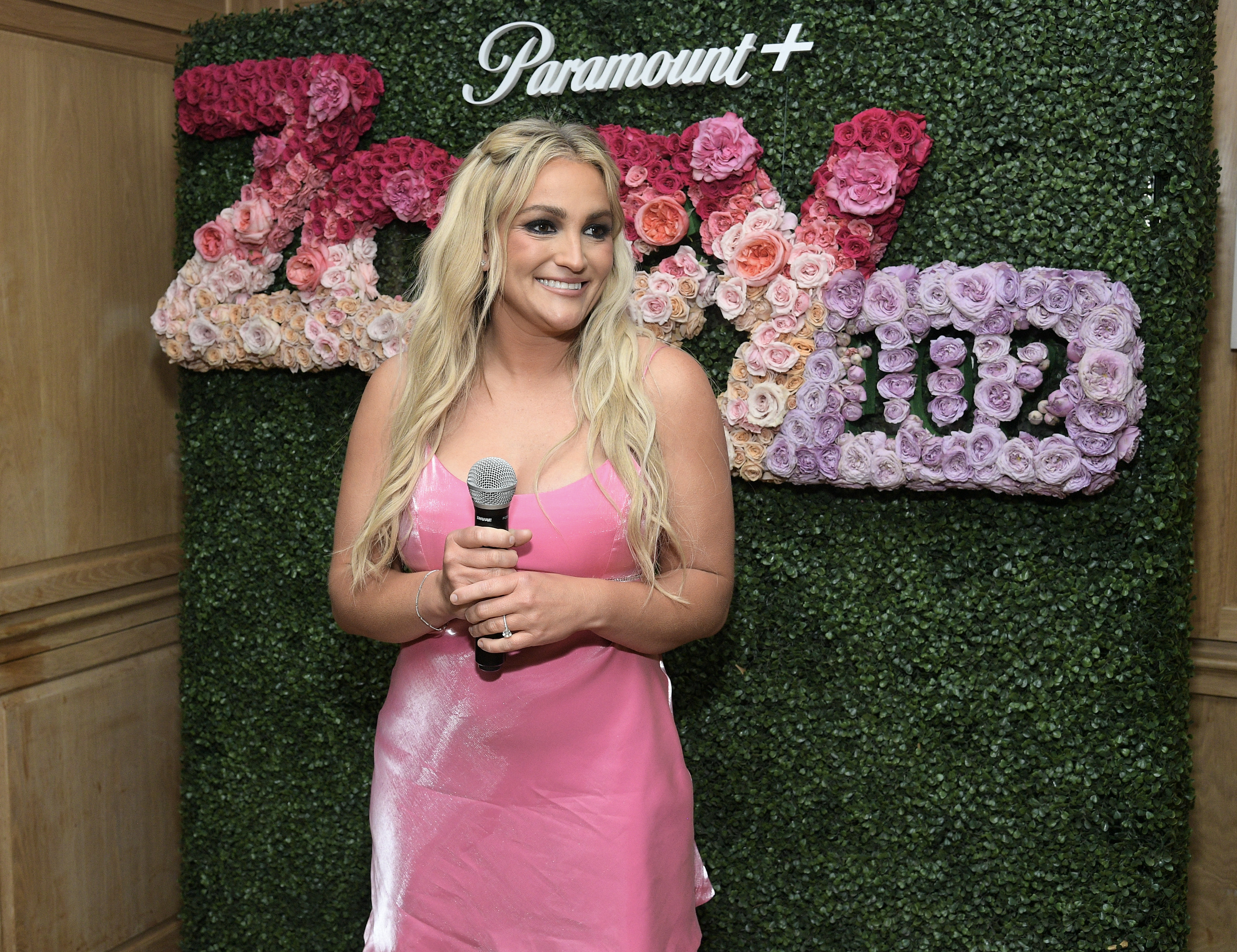 Jamie Lynn Spears, in a sleeveless dress, holds a microphone standing in front of a 'Zoey 102' flower backdrop at a Paramount+ event