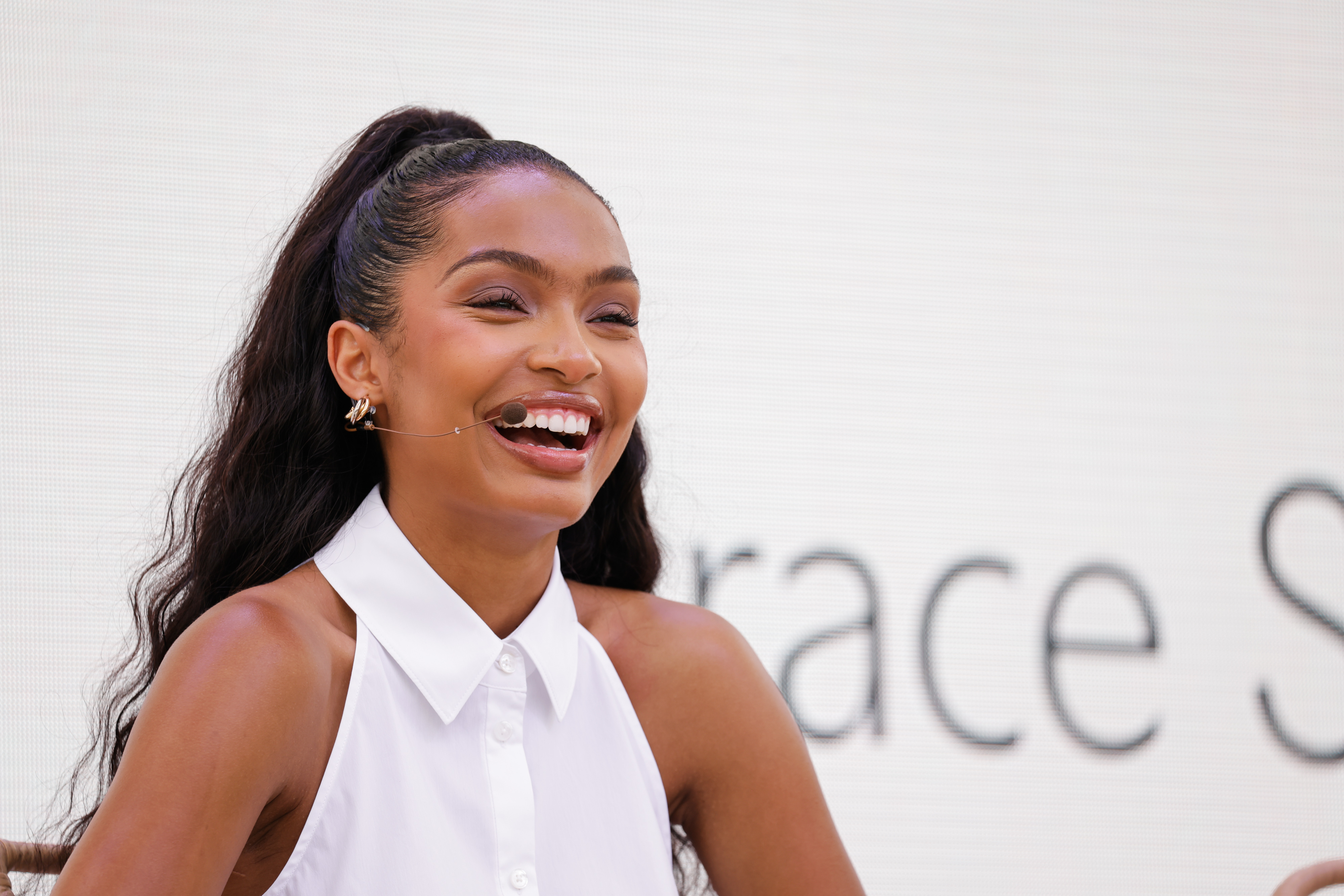 Yara Shahidi smiles while speaking at an event, wearing a sleeveless white blouse with a high ponytail