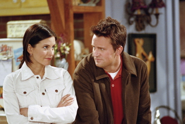 Courteney Cox and Matthew Perry in a scene from the TV show Friends, with Courteney in a white jacket and Matthew in a brown jacket over a red shirt