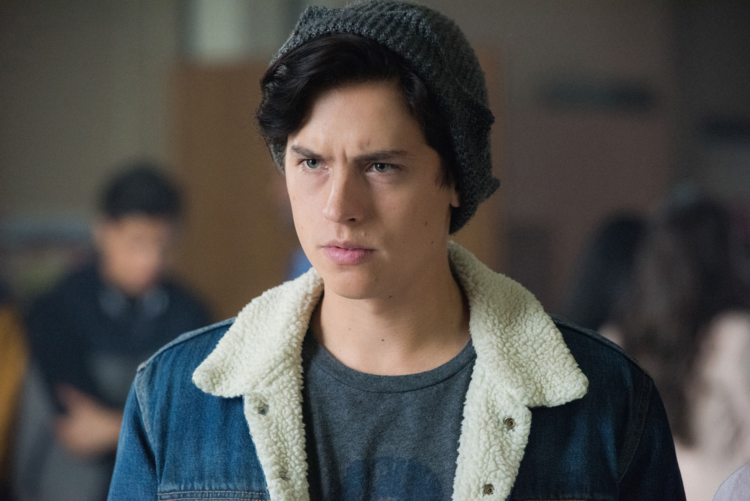 Cole Sprouse, wearing a denim jacket with shearling lining and a grey beanie, portrays Jughead Jones in a scene from the TV show "Riverdale."