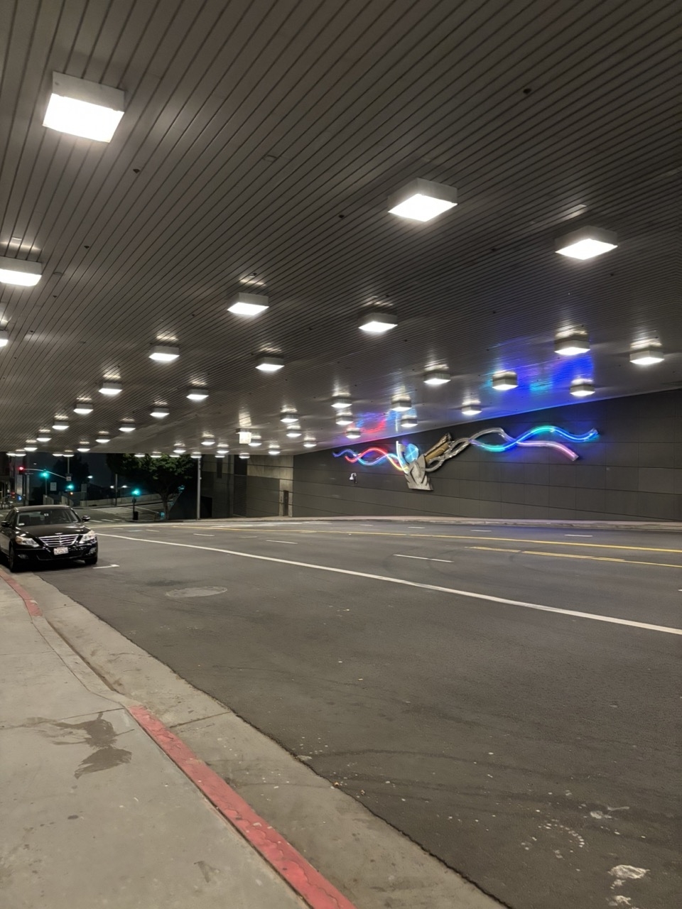 An urban street scene at night, featuring an empty road with a car parked on the side. Neon lights on the building's exterior display a wave-like design