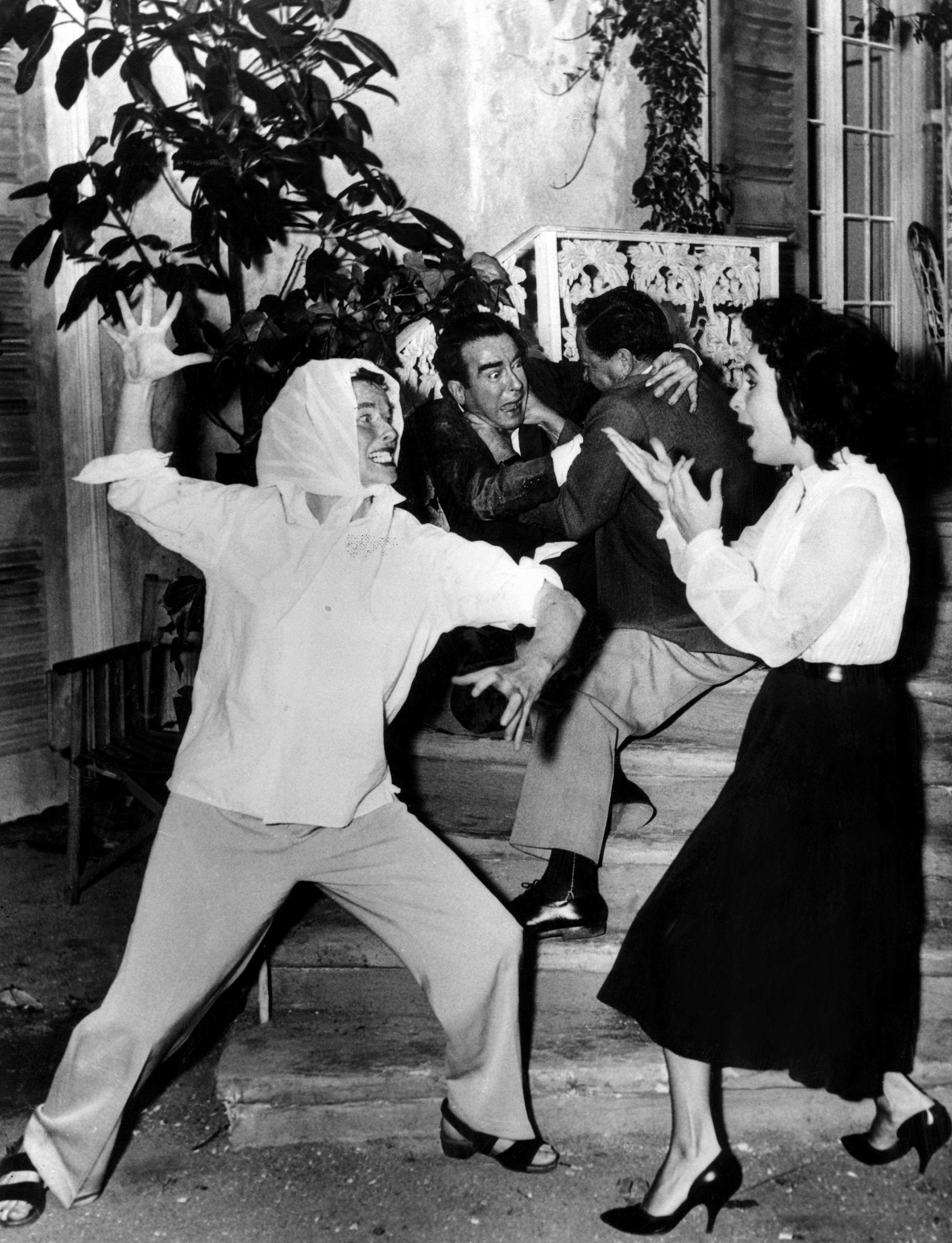 A behind-the-scenes, black-and-white shot of Elizabeth Taylor, Richard Burton, and Rex Harrison playfully acting during a film's production