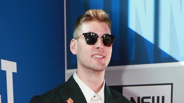 A man on the red carpet wearing sunglasses, a white shirt, and a dark blazer with a pin