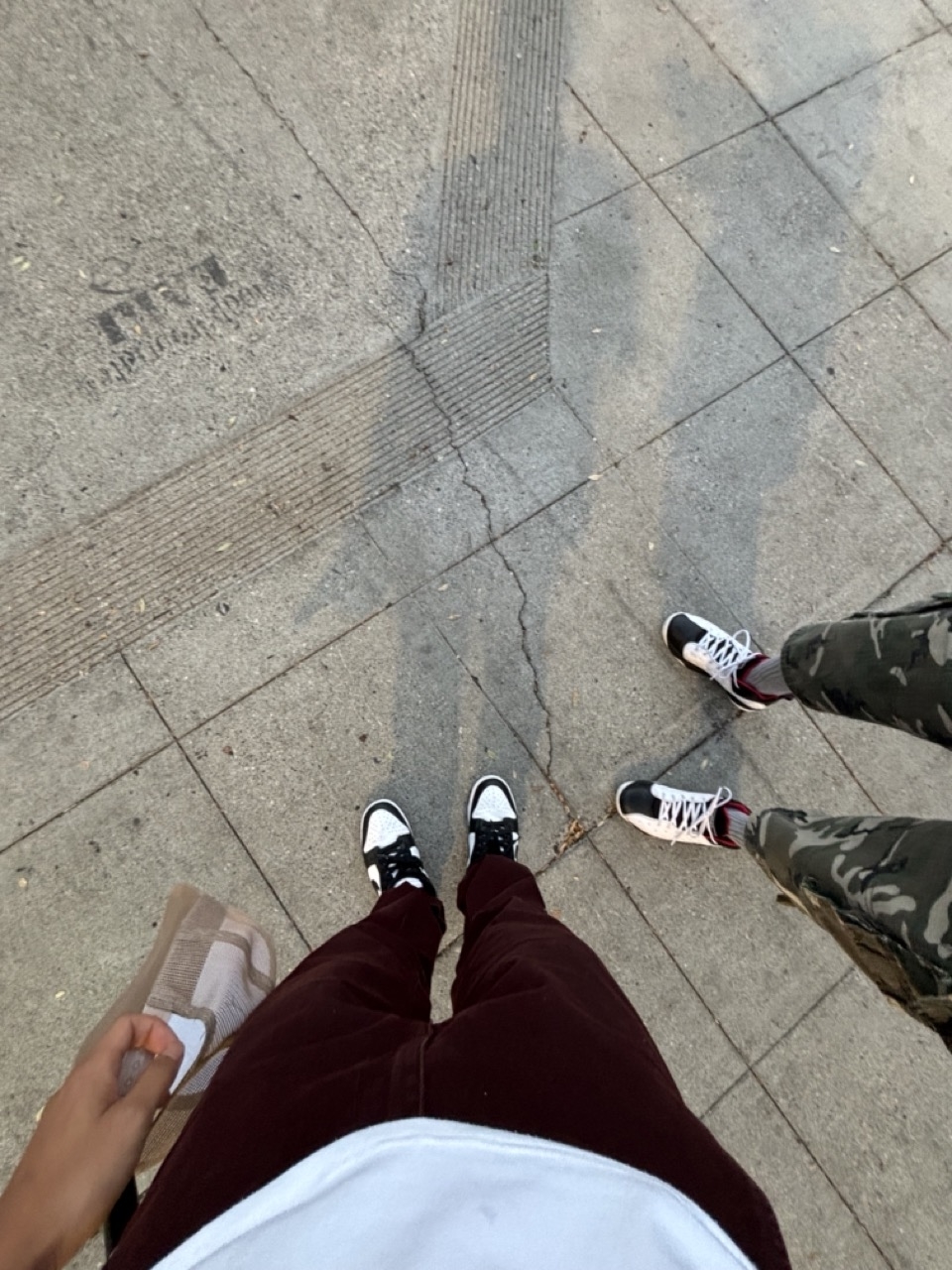Two people standing on a pavement; one wearing maroon pants, black and white shoes, and holding a brown bag; the other in camouflage pants and black and white shoes