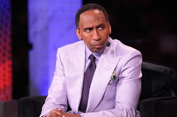 Stephen A. Smith in a light-colored blazer and tie sits on stage, looking focused