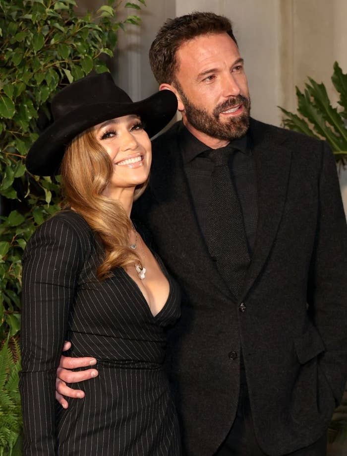 Jennifer Lopez in a pinstripe suit dress and black hat, smiles alongside Ben Affleck in a black suit and tie, posing for a photo together