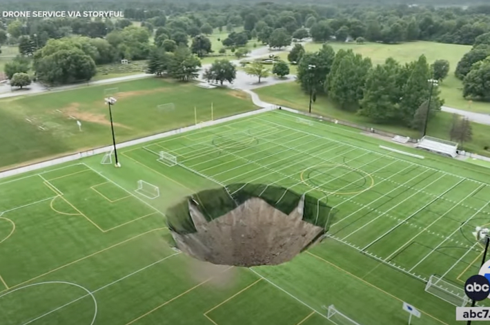 A drone photo shows a large sinkhole in the middle of an athletic field with soccer and lacrosse markings, reported by ABC 7 News