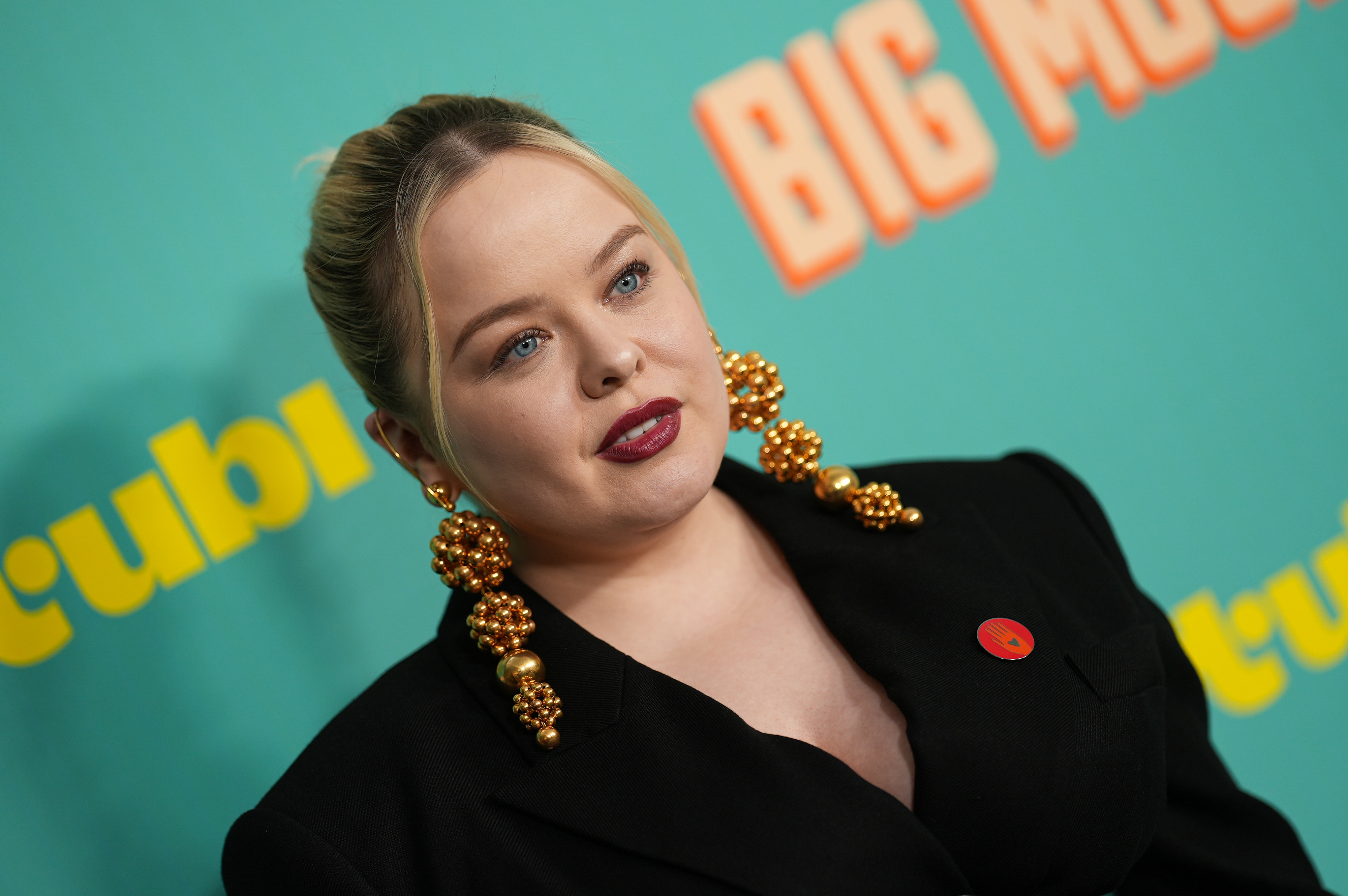 Nicola Coughlan attending an event, wearing a black outfit with large, golden beaded earrings, standing in front of a backdrop that reads "BIG MOVIE" and "tubi"