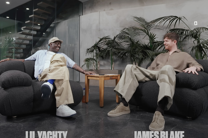Lil Yachty and James Blake sit on black armchairs facing each other, smiling and talking in a modern setting with a wooden table and plants in the background
