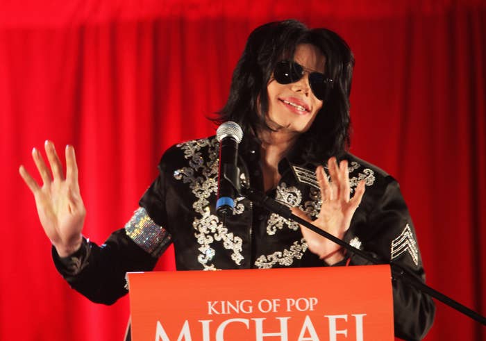 Michael Jackson, wearing a black embellished jacket and sunglasses, speaks at a podium with &quot;King of Pop Michael Jackson&quot; written on it