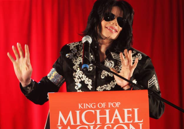 Michael Jackson, wearing a black embellished jacket and sunglasses, speaks at a podium with "King of Pop Michael Jackson" written on it