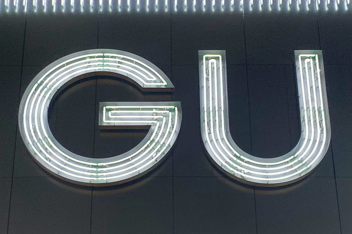 The image features an illuminated store sign reading &ldquo;GU&rdquo; against a dark backdrop with vertical light strips surrounding it