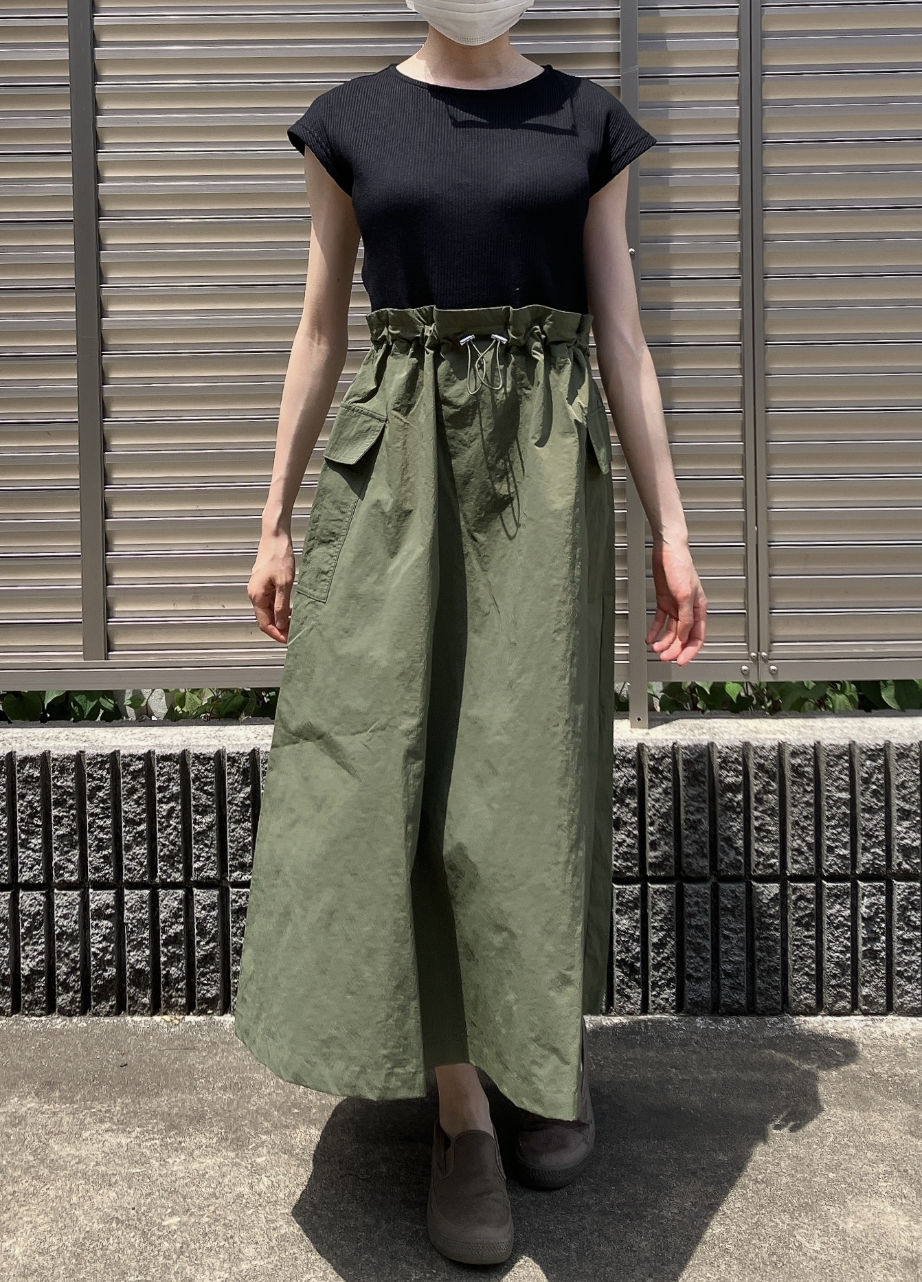 A person wearing a black top, green high-waisted skirt with drawstrings, and brown shoes stands outside. Their face is covered with a mask