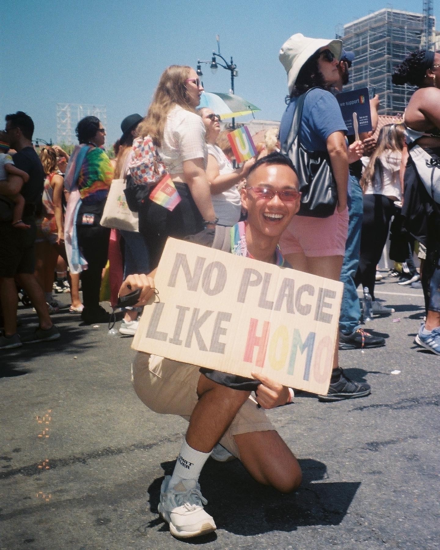 Person crouching at a Pride event, holding a sign that reads "No Place Like Homo," smiling, and surrounded by other attendees
