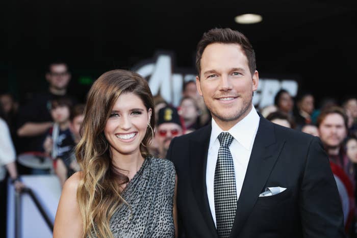Chris Pratt and Katherine Schwarzenegger smile on the red carpet. Chris wears a suit and tie, and Katherine wears a one-shoulder gown. Fans in the background