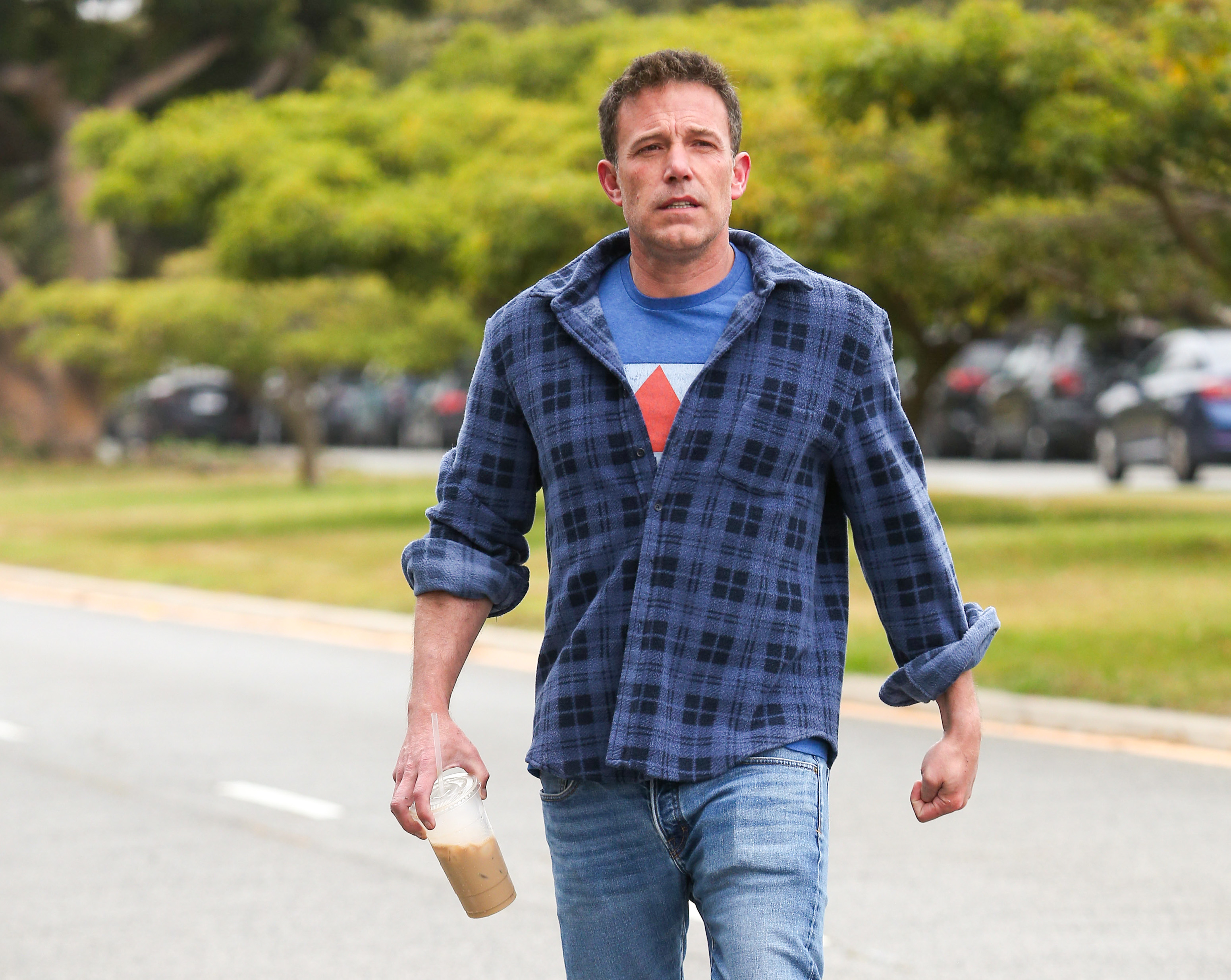 Ben Affleck walks outdoors holding an iced coffee, wearing a blue checkered shirt over a blue t-shirt with a star on it and jeans