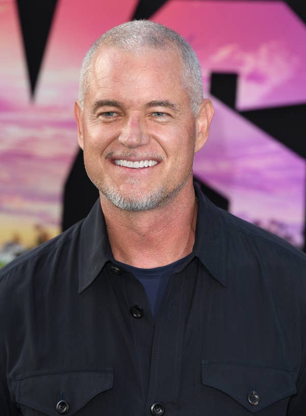 Eric Dane is smiling at a formal event, wearing a dark-colored button-up shirt with a crew neck t-shirt underneath. The background shows a colorful abstract pattern