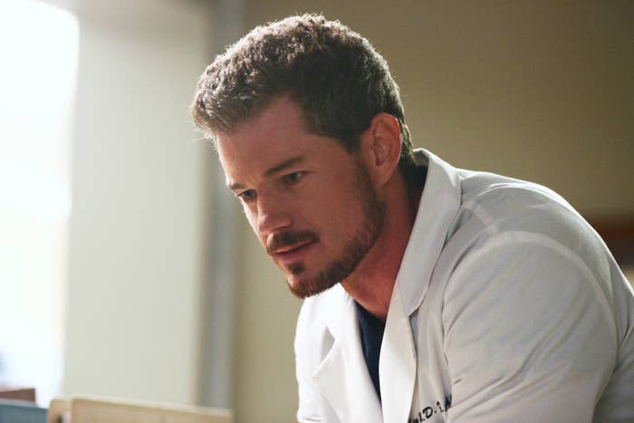 Eric Dane, in a white lab coat, looking down thoughtfully. The background is indistinct
