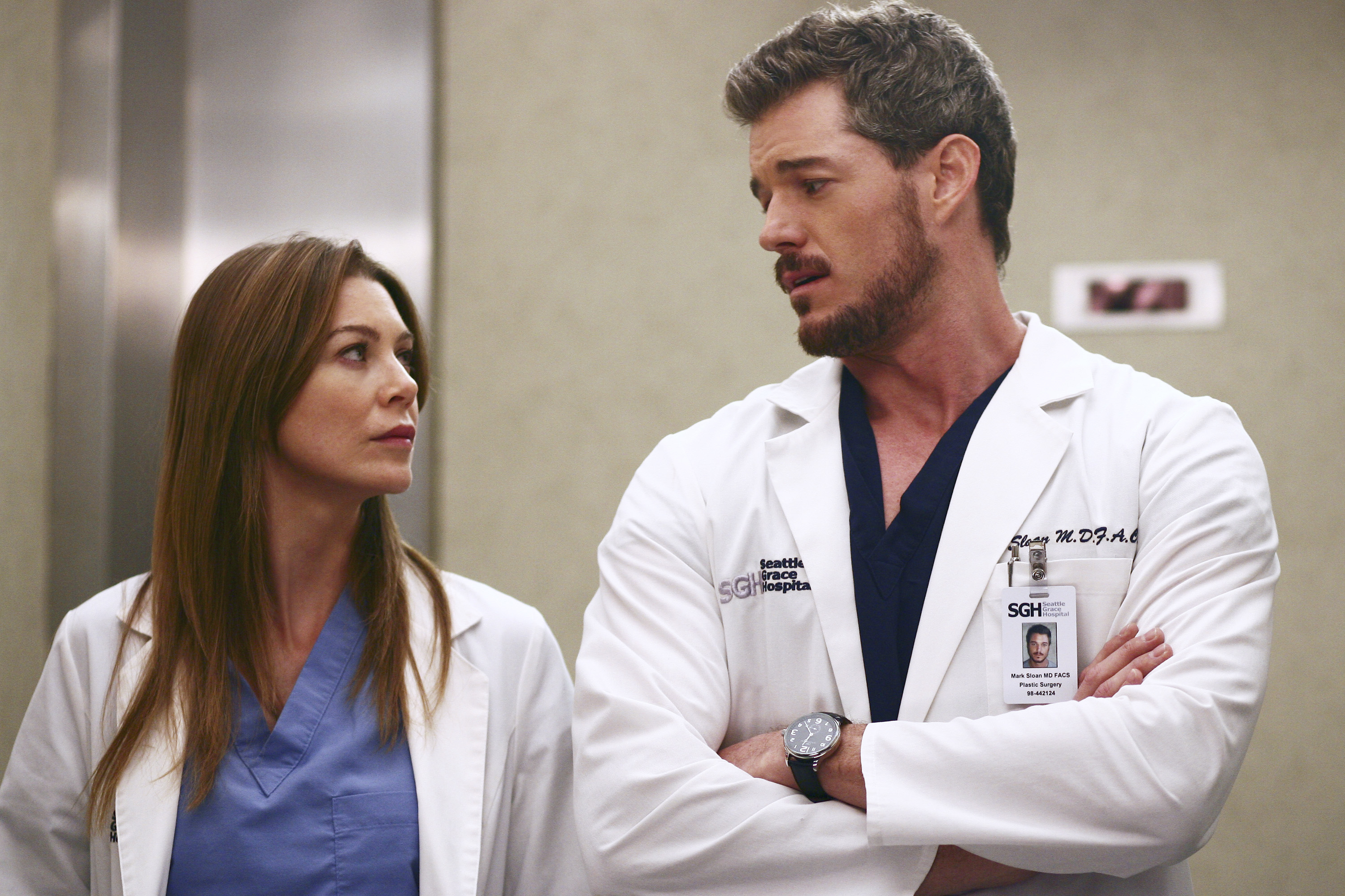 Ellen Pompeo and Eric Dane in hospital scrubs and white coats, appearing to be in a medical setting, looking at each other with serious expressions