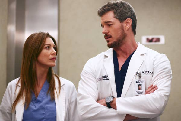 Ellen Pompeo and Eric Dane in hospital scrubs and white coats, appearing to be in a medical setting, looking at each other with serious expressions