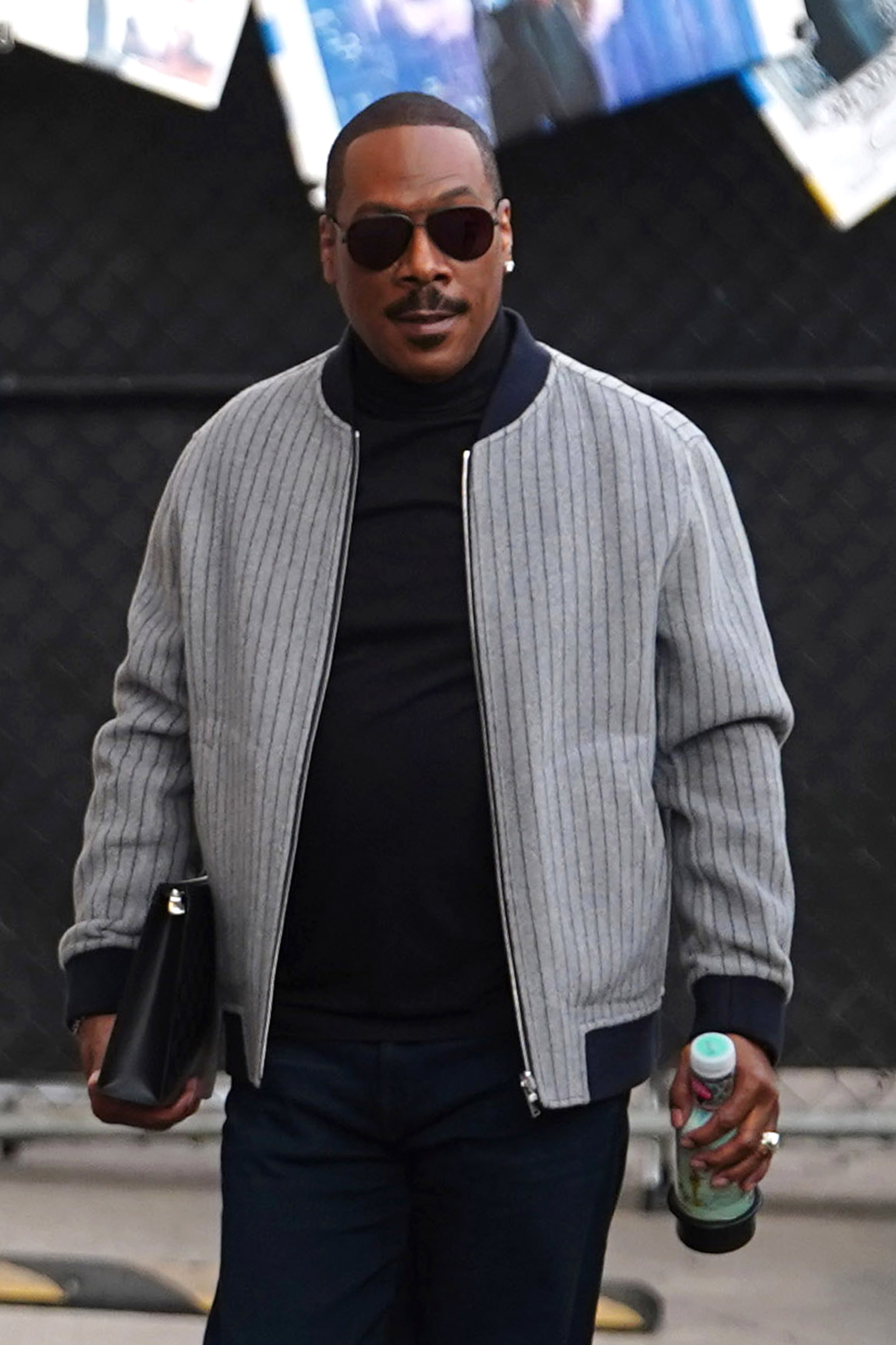 Eddie Murphy wearing sunglasses, holding a water bottle and a black folder, dressed in a striped jacket and dark pants
