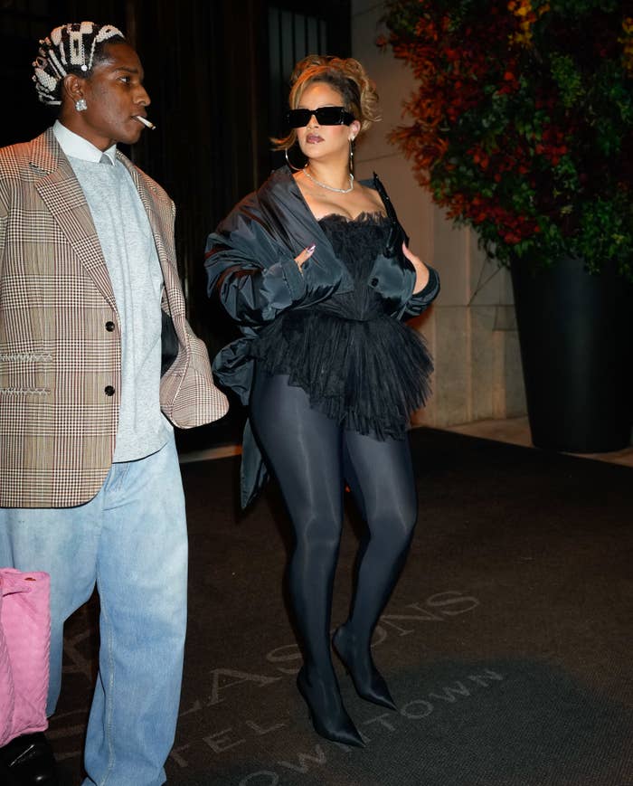 Rihanna in a sheer black dress, tights, and high heels, standing with A$AP Rocky who is wearing a plaid jacket and loose jeans with a white beanie and hair clips