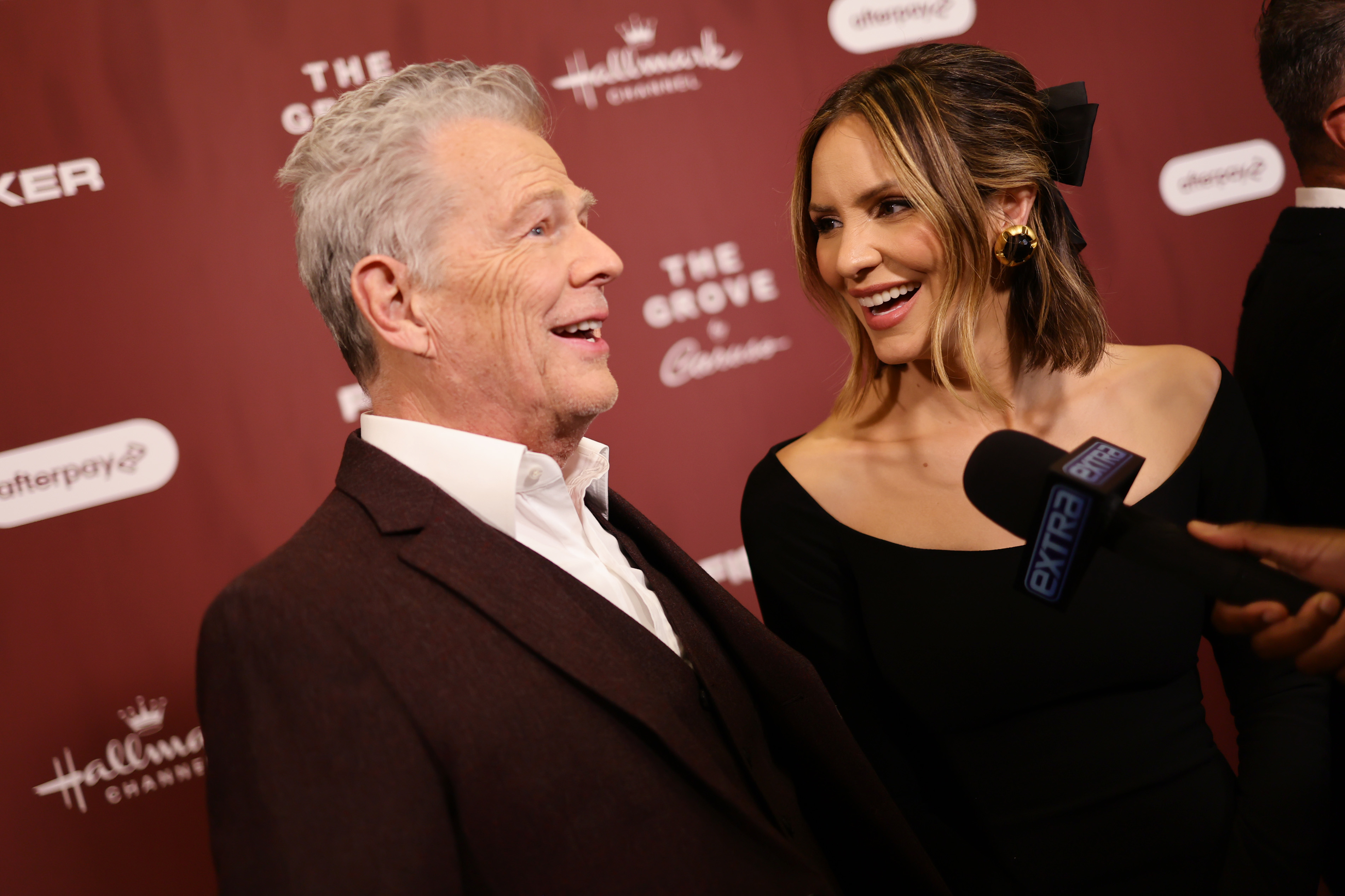David Foster and Katharine McPhee are smiling on the red carpet, with Katharine wearing an off-shoulder outfit and a black bow in her hair