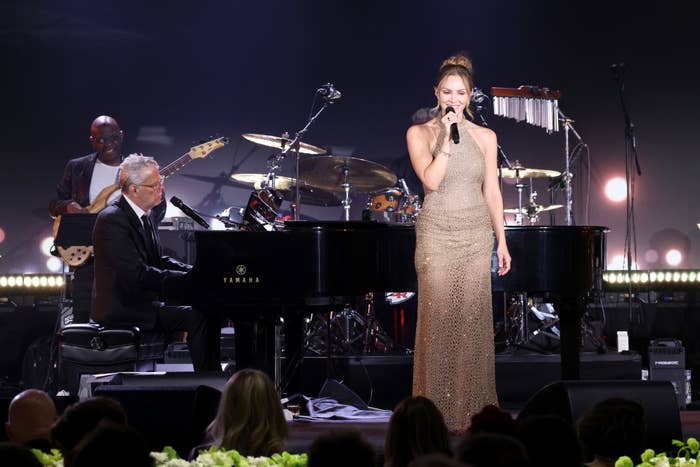 David Foster playing the piano while Katharine McPhee sings on stage in a sparkling gown at a concert. A guitarist and drummer are seen in the background