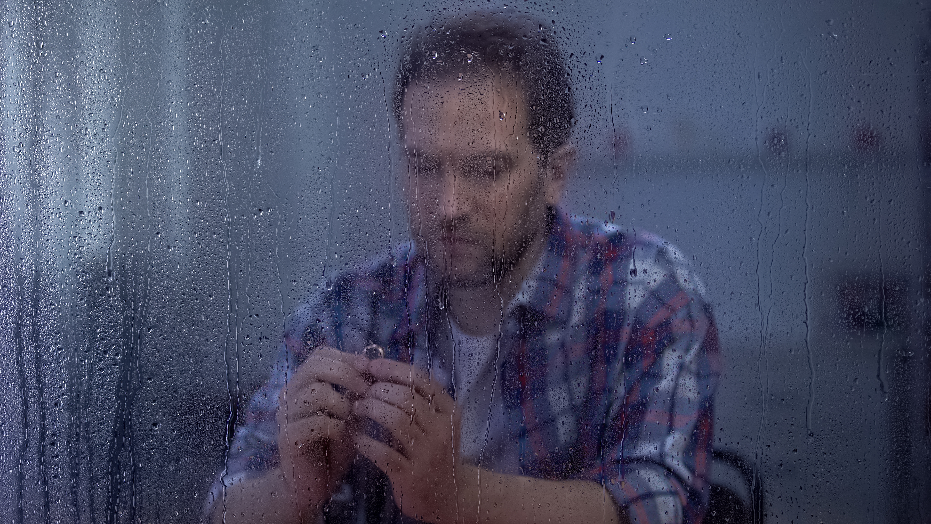 A man, blurred by a rainy window, sits thoughtfully, holding a ring in his hands, conveying a sense of contemplation or emotional reflection