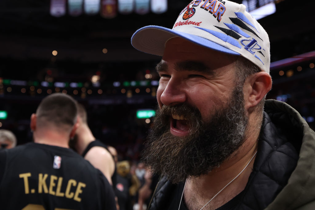 Jason Kelce wears a casual jacket and a sports cap with the word "All-Star" on it, smiling at an event. Travis Kelce is nearby in a jersey