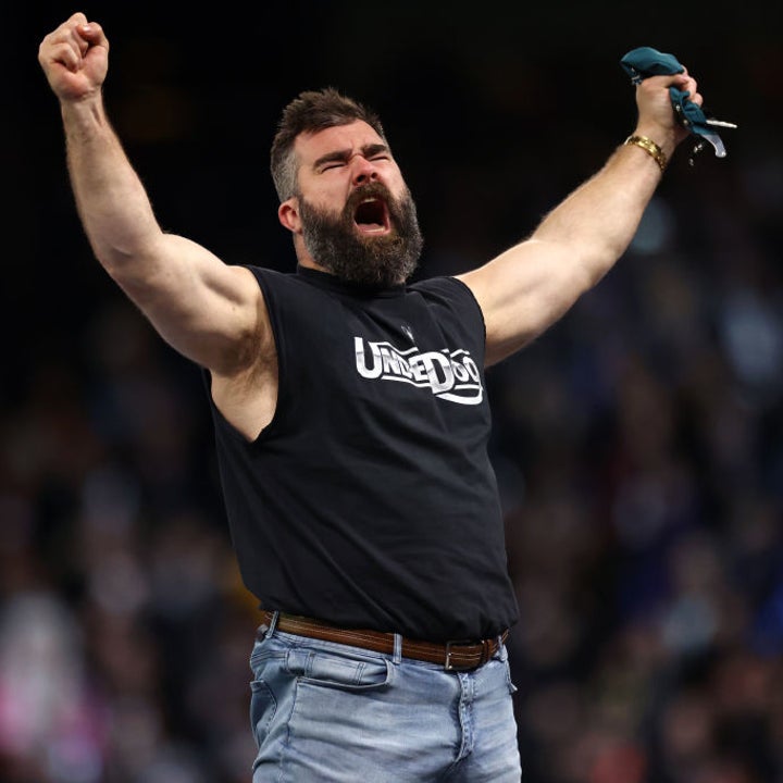 Jason Kelce cheers with arms raised, wearing a sleeveless "Underdog" shirt and jeans, in a lively crowd