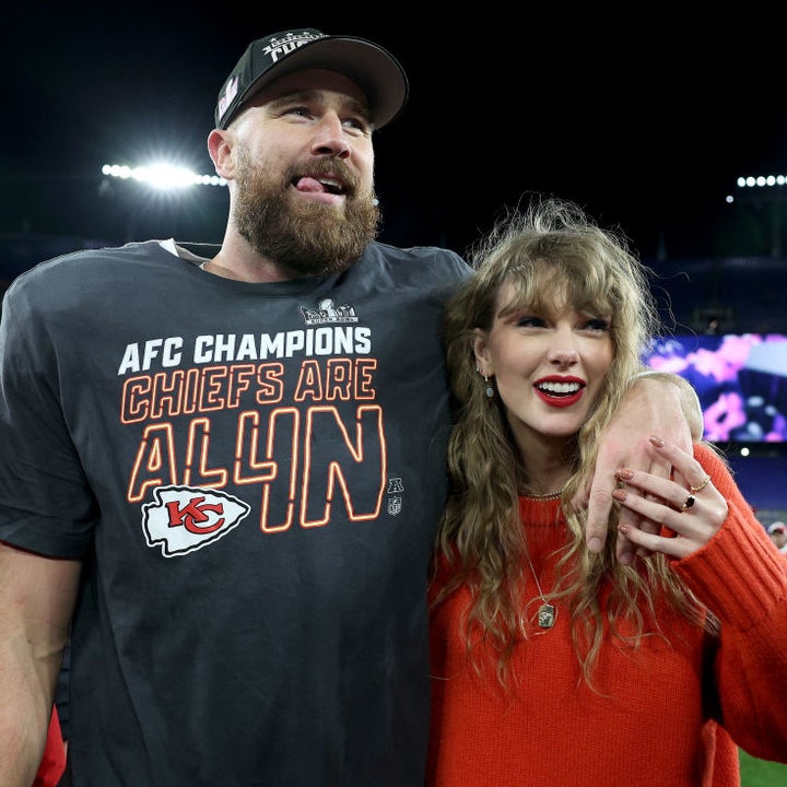 Travis Kelce in a "AFC Champions Chiefs Are All In" shirt with Taylor Swift in a red sweater, smiling and posing together on a sports field