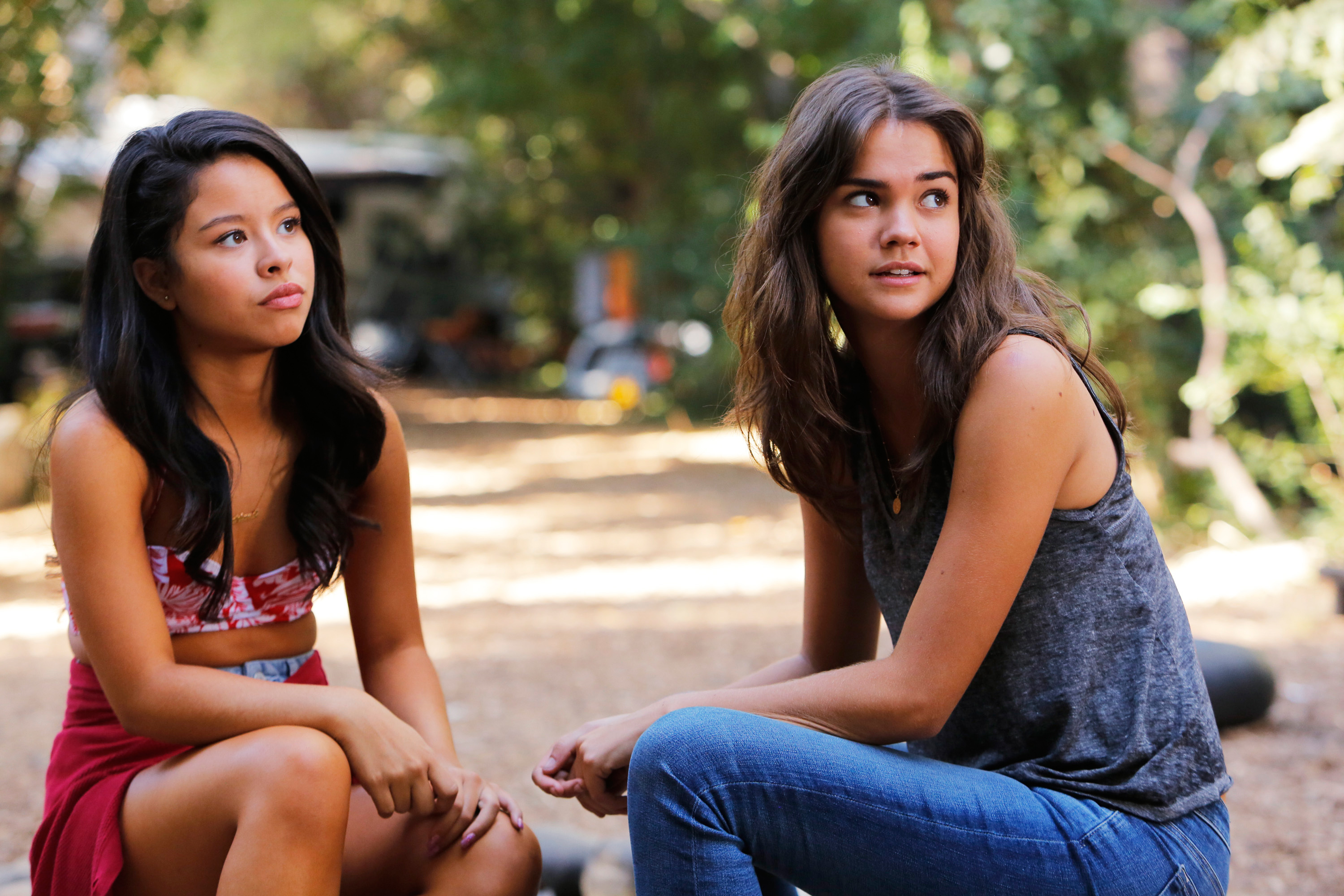 Cierra Ramirez and Maia Mitchell are seated outdoors, engaging in conversation. Cierra wears a cropped top and skirt; Maia wears a sleeveless top and jeans