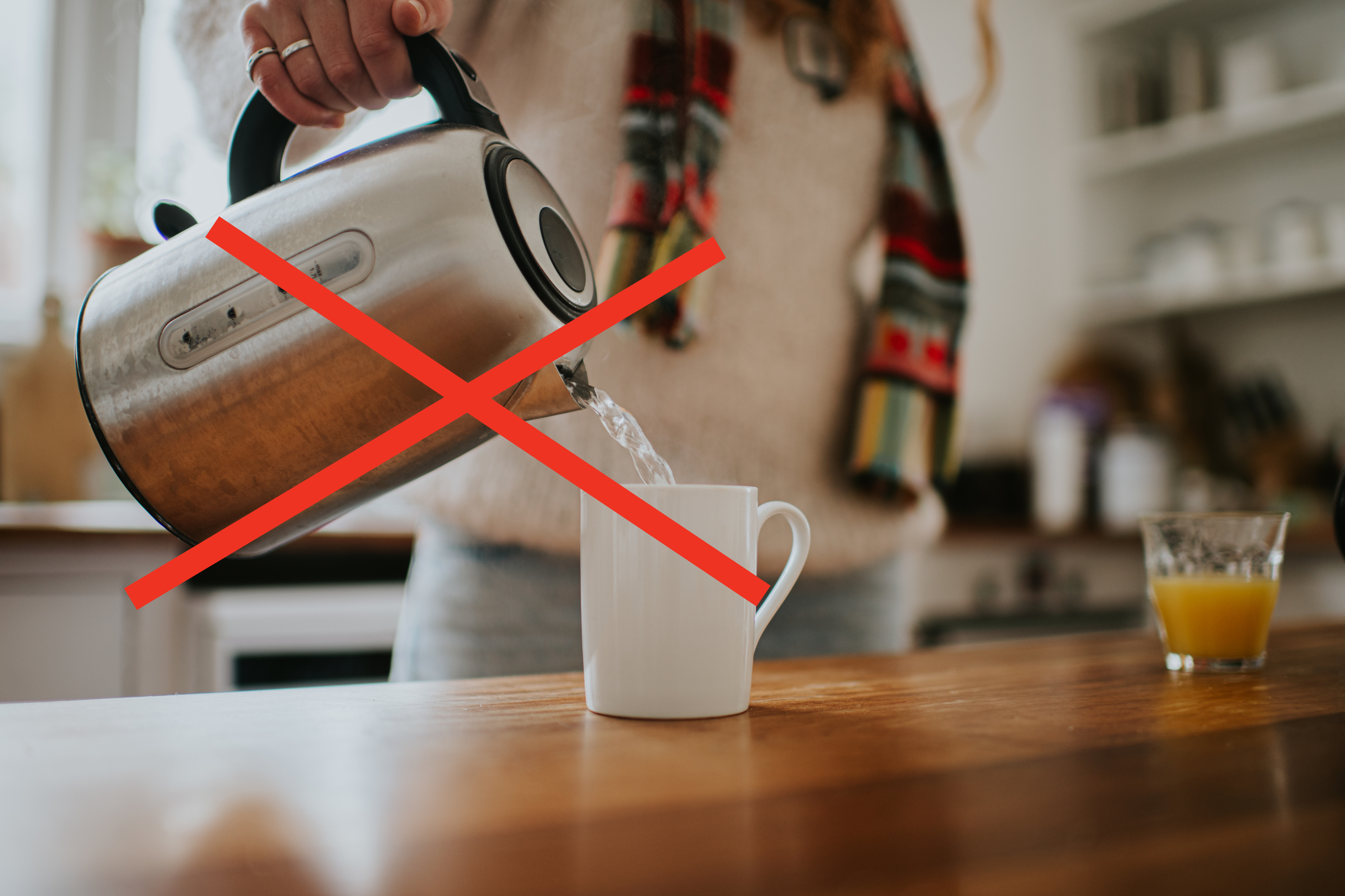 A person pours hot water from a kettle into a white mug on a kitchen counter. A glass of orange juice is nearby