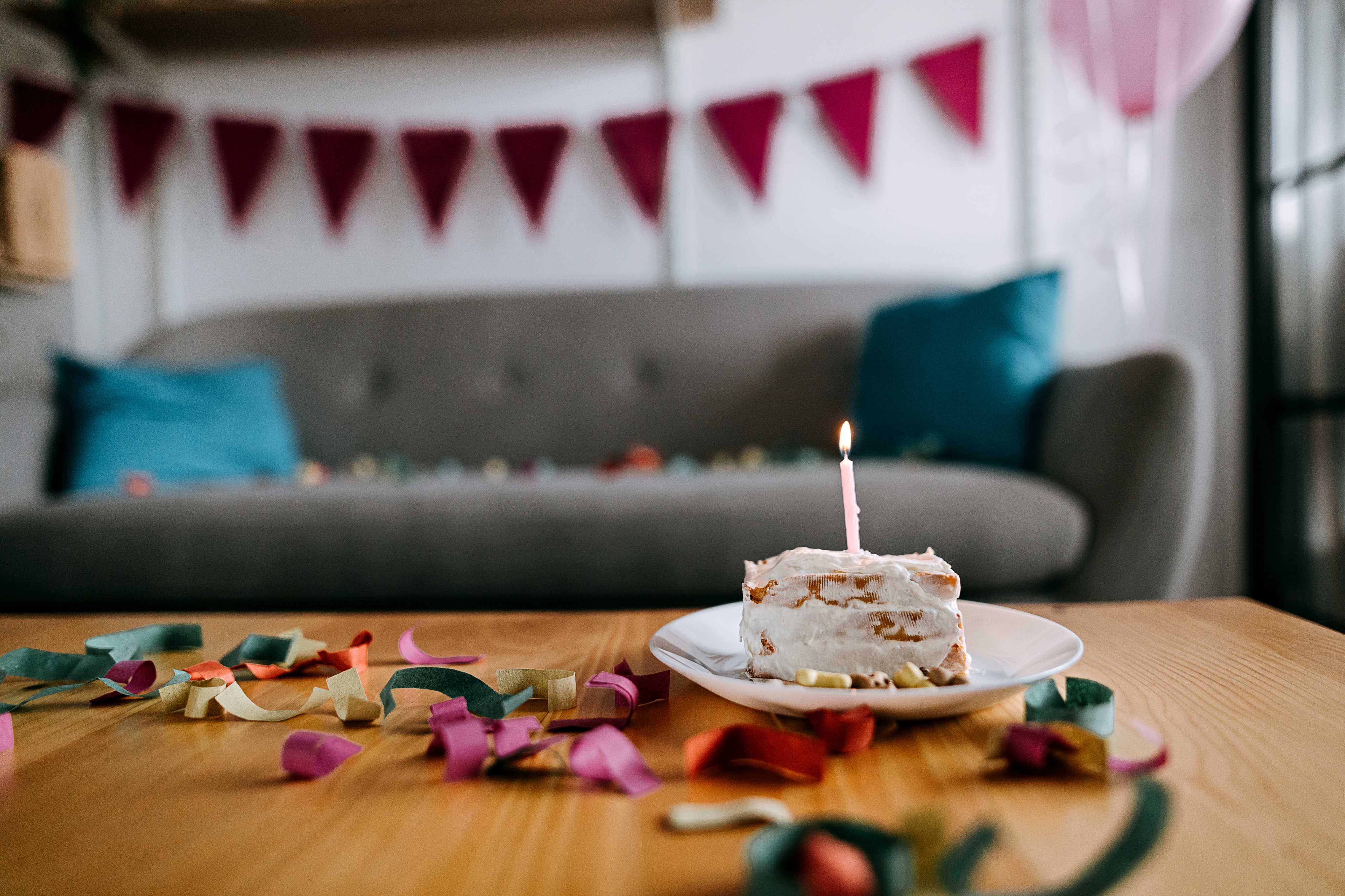 Slice of cake with a lit candle on a table, surrounded by colorful confetti, with a couch and party decorations in the background