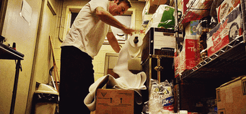 A man in a storage room humorously struggling to control a large roll of paper towels that are unwinding and falling off a shelf