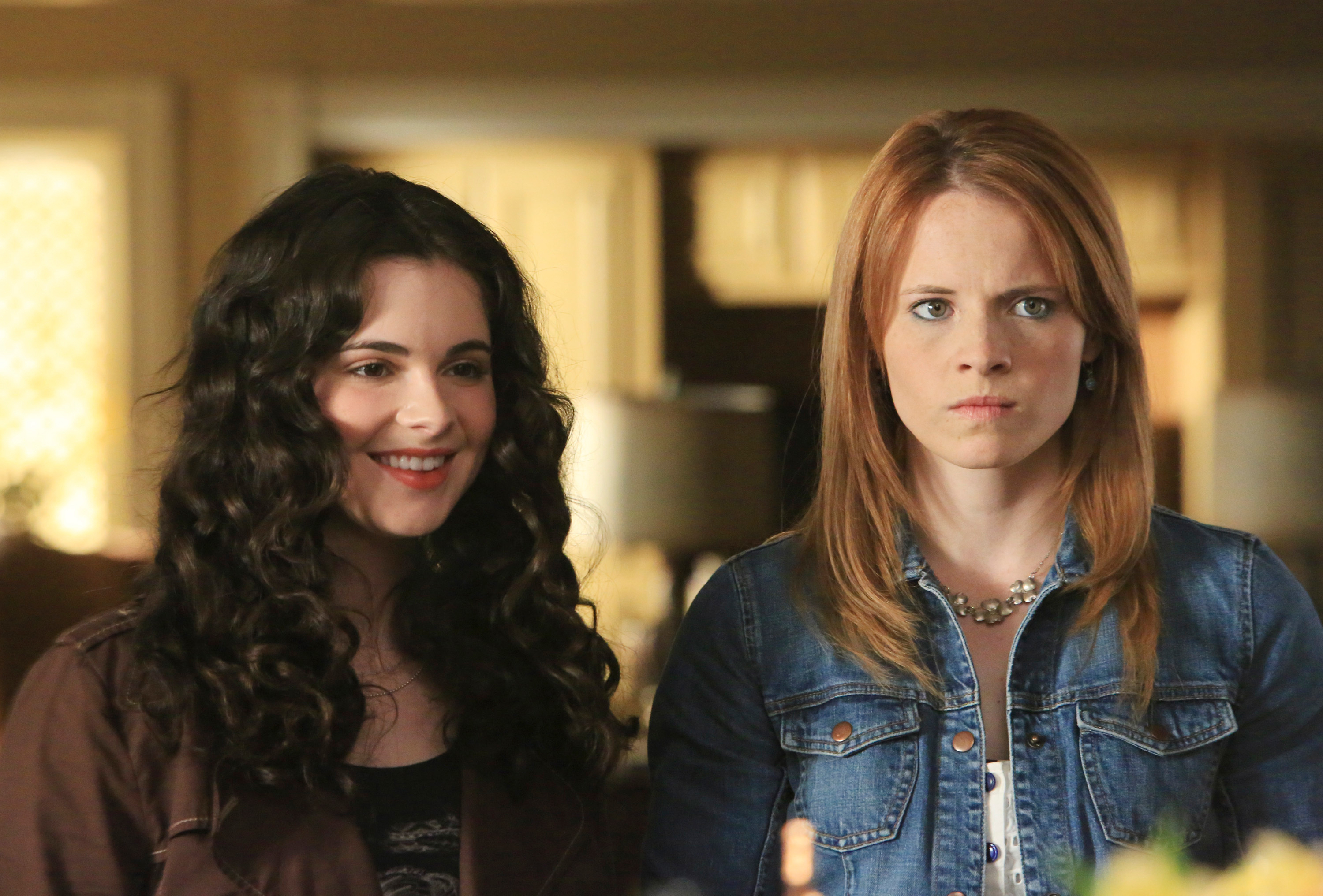 Vanessa Marano and Katie Leclerc stand side by side; Vanessa smiles, wearing curly hair, Katie appears serious in a denim jacket with straight hair