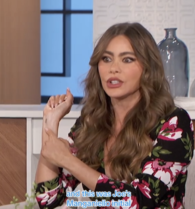 Sofía Vergara, seated, gestures while discussing Joe Manganiello's initial tattoo. She is wearing a floral print outfit