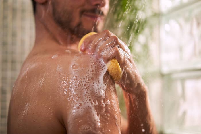 A shirtless man is taking a shower, using a yellow sponge to scrub his shoulder. His face is partially visible and he is smiling