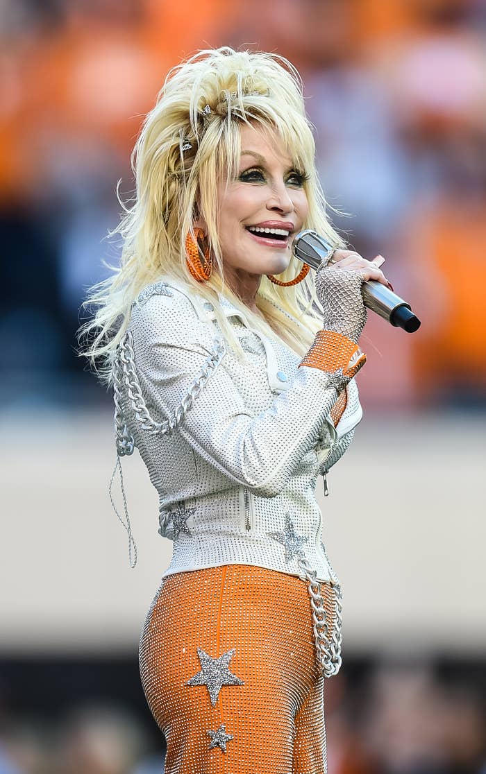Dolly Parton sings on stage wearing a sparkling outfit adorned with stars, holding a microphone close to her mouth, and smiling