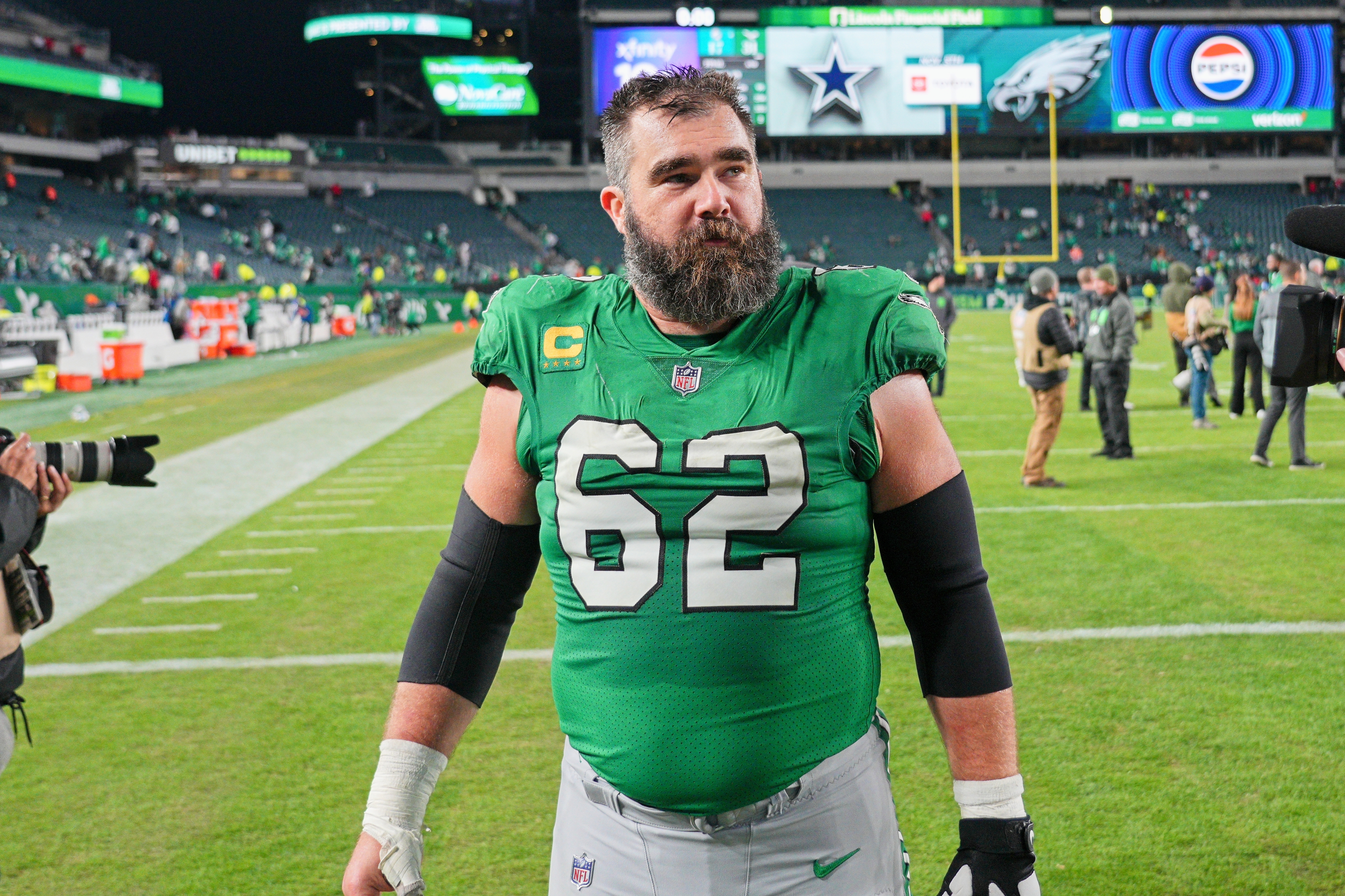 Jason Kelce on a football field wearing his green team uniform with the number 62, arm sleeves, and a black glove, looking towards the camera