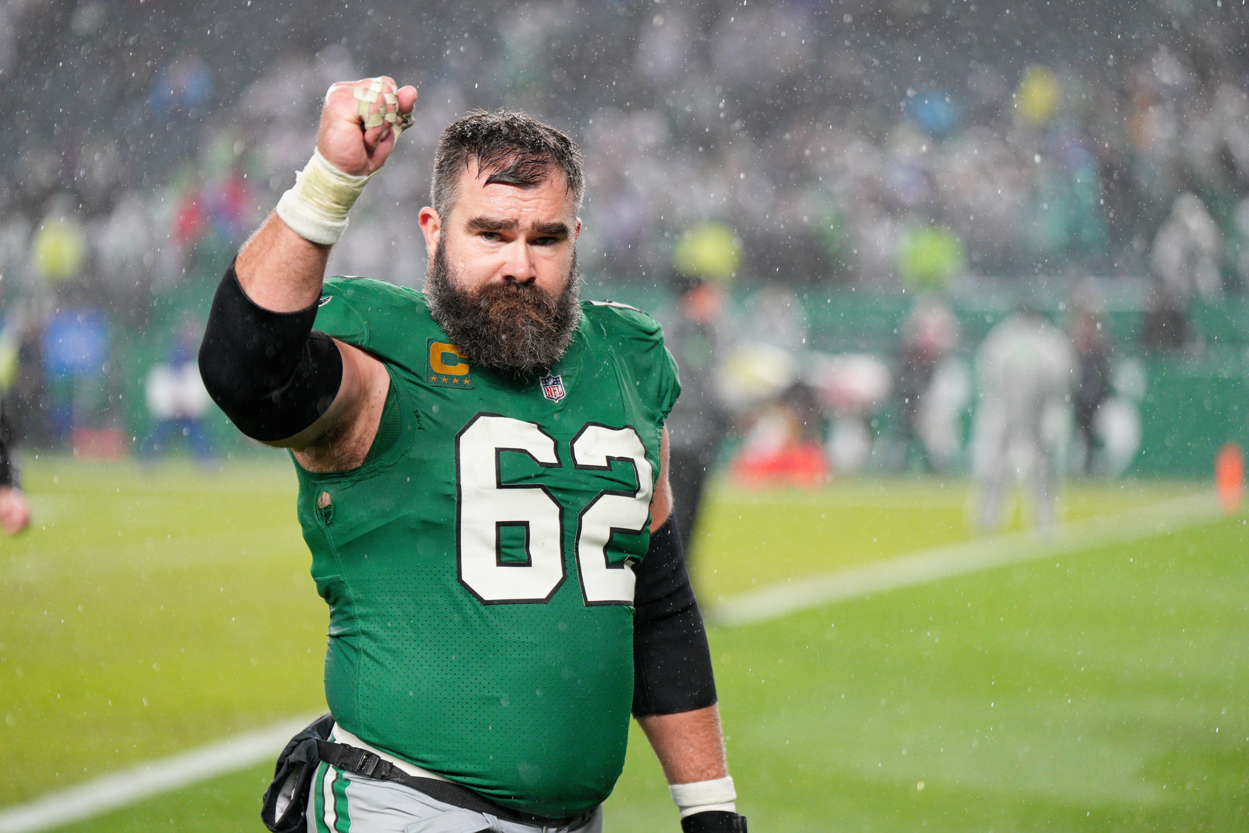 Jason Kelce, wearing a green football uniform with the number 62, raises his fist with a determined expression on the field during a rainy game