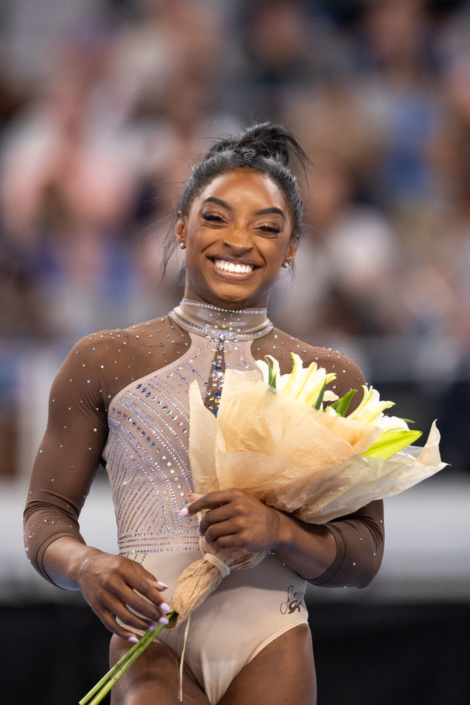 Simone Biles smiles and holds a bouquet of flowers while dressed in a sparkling gymnastics leotard