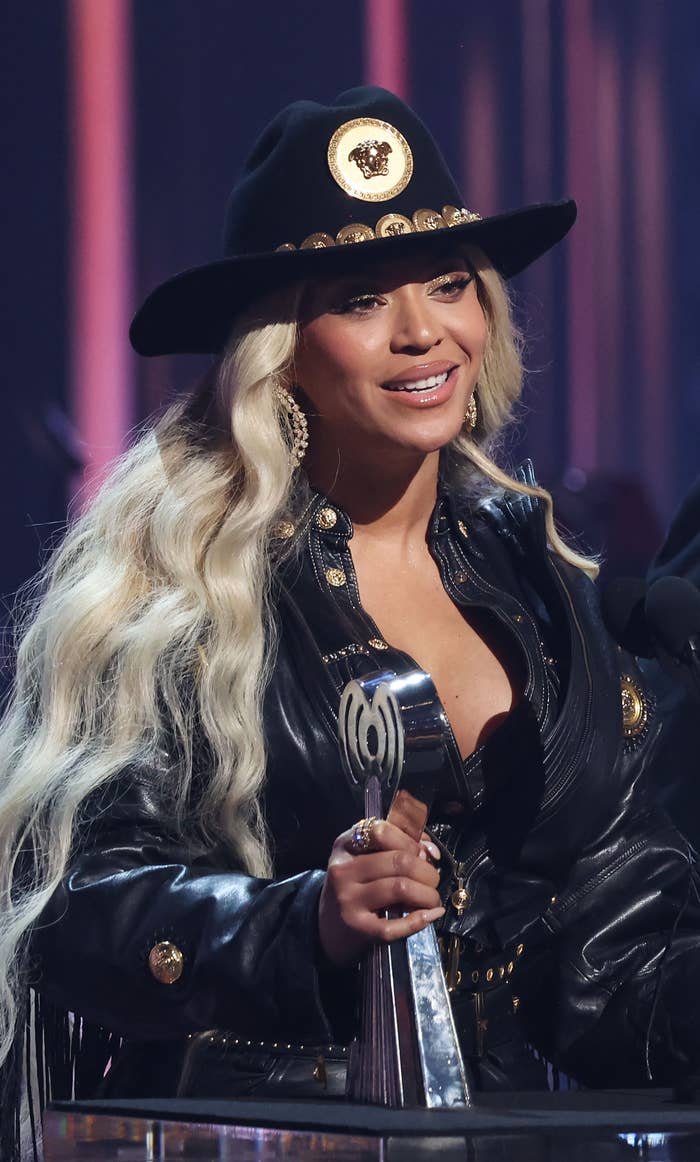 Beyoncé smiling while holding a trophy and wearing a cowboy hat and a stylish leather jacket with gold embellishments, speaking at an event